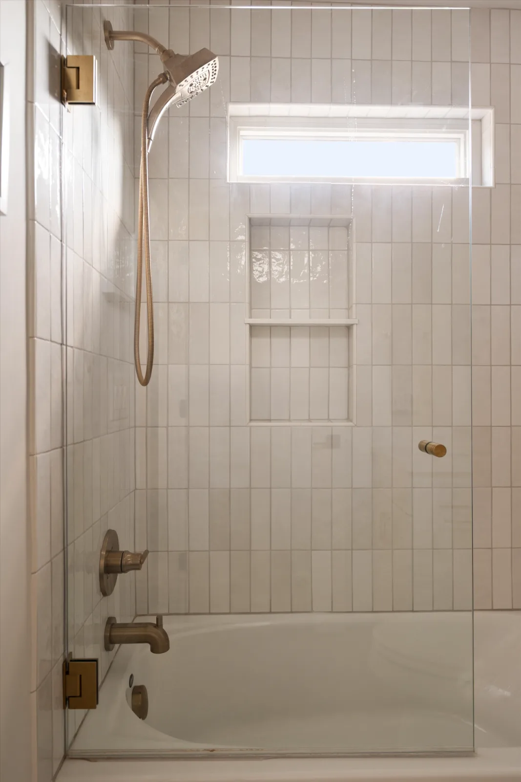 The tub and shower: vertical white tile, a recessed niche, a clerestory window and brass fittings